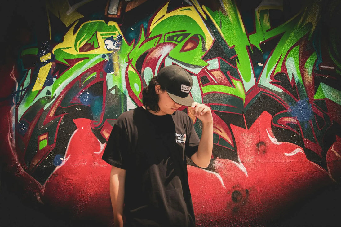A young man wearing a black cap stands against a colorful graffiti wall, creating a striking urban scene.