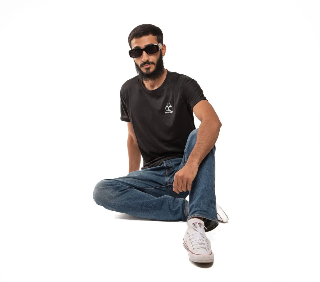 A stylish young man in a black shirt and jeans, seated against a white background.
